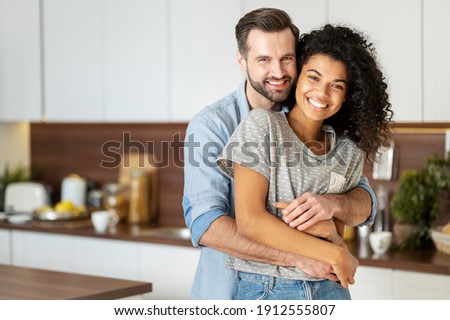 Smiling man hugging from behind charming African American woman, two people standing and joyfully looking at camera. Young international couple happily spending time in cozy modern kitchen at home. Royalty-Free Stock Photo #1912555807