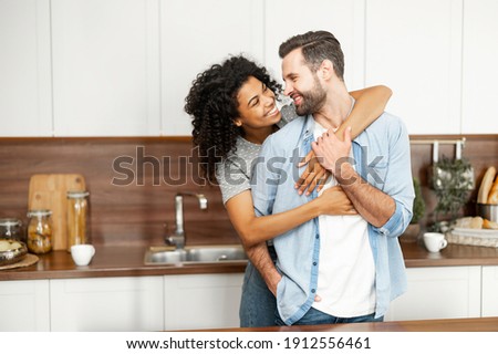 Close-up of a happy interracial couple standing in the kitchen, happy romantic owners of a new flat smiling and looking at each other, young African American woman hugging handsome man from behind Royalty-Free Stock Photo #1912556461