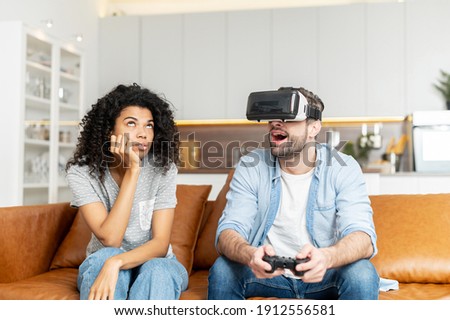 Interracial couple sitting on the couch, addicted guy in vr headset and with joystick is playing video game without paying attention to young African American girlfriend, she is bored and annoyed