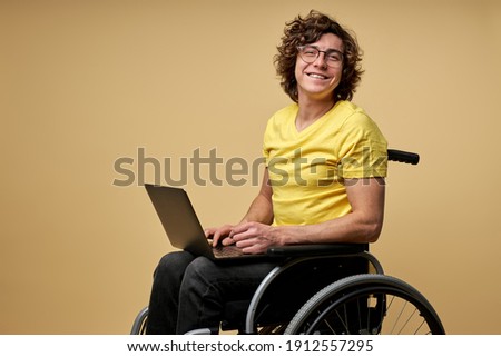 disabled man sits in a wheelchair with laptop, working online. curly young male in casual wear smiles at camera isolated in studio on beige background Royalty-Free Stock Photo #1912557295