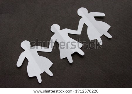 Figures of three girls in dresses holding hands, cut out of white paper. In the center of the photo on a black background