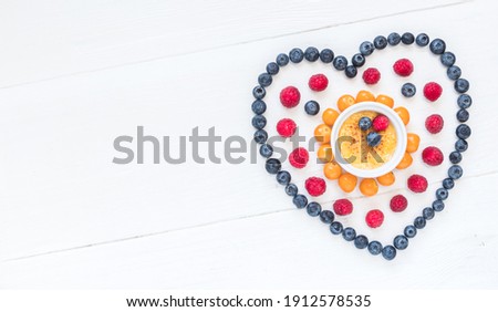 Heart symbol made of different berries with a creme brulee in the middle on white wood, top view with copy space