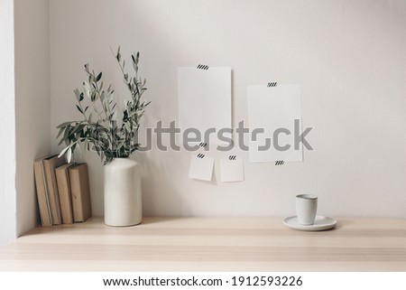 Breakfast still life. Cup of coffee, books on wooden desk, table. Empty notepads and posters mockups taped on white wall.Vase with olive branches. Elegant Scandinavian working space, home office. Royalty-Free Stock Photo #1912593226