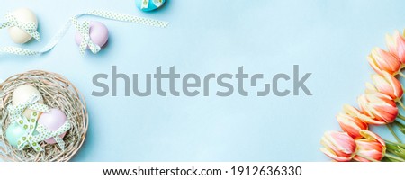 Golden easter colour eggs in basket with spring tulips, white feathers on pastel blue background in Happy Easter decoration. Spring holiday top view banner
