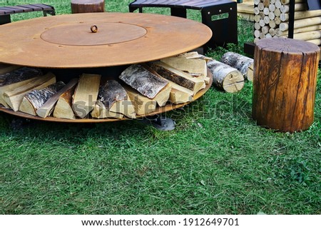 Round Patio Iron Fire Pit Table On The Grass For Outdoor Leisure Party. Steel Rounded Fire Pit With Grill Top On Backyard Party Place. Grill Appliance And Fireplace On The Back Yard Lawn.