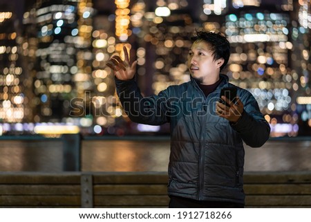 Asian man wearing overcoat suit using smart mobile phone with smile action over the photo blurred bokeh of New york Cityscape beside the east river, USA downtown, career working everywhere concept