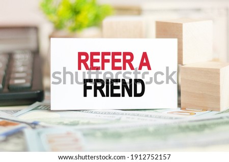 The words REFER A FRIEND is written on white paper card near a wooden cubes, calculator on a banknotes background. Business and Financial concept