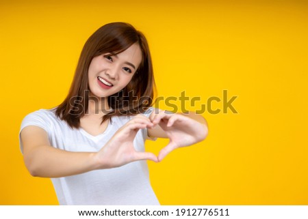 Young elegant Asian woman smiling and make heart sign by hands isolated on yellow background