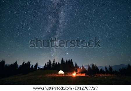 Silhouette of male traveler near illuminated camp tent under beautiful night sky with stars. Magnificent view of blue starry sky under grassy hill. Concept of travelling, hiking and night camping.