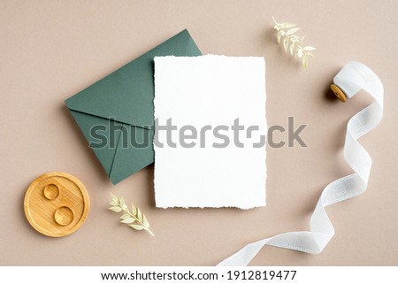 Wedding invitation card mockup, green envelope, golden rings, silk ribbon, dried flowers on pastel beige background. Flat lay, top view, copy space. Rustic wedding stationery set.