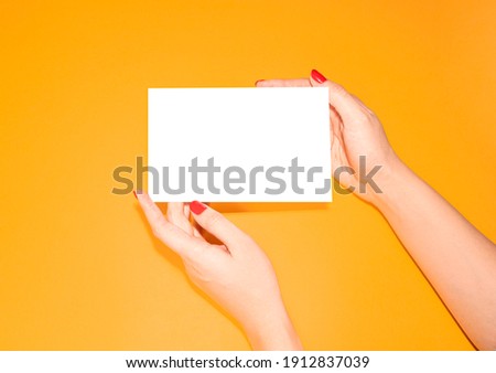 Female hands with beautiful red manicure hold a discount card, business card, mockup of a blank white sheet of paper on a yellow background with copy space. Template for design yellow background. 