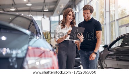 Positive young professional saleswoman demonstrating document to male customer buying new car in dealership Royalty-Free Stock Photo #1912865050