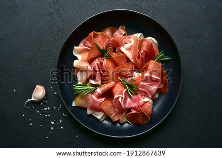 Slices of prosciutto di parma or jamon serrano (iberico)  on a black plate on a dark slate, stone or concrete background. Top view with copy space. Royalty-Free Stock Photo #1912867639