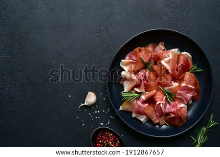 Slices of prosciutto di parma or jamon serrano (iberico)  on a black plate on a dark slate, stone or concrete background. Top view with copy space. Royalty-Free Stock Photo #1912867657