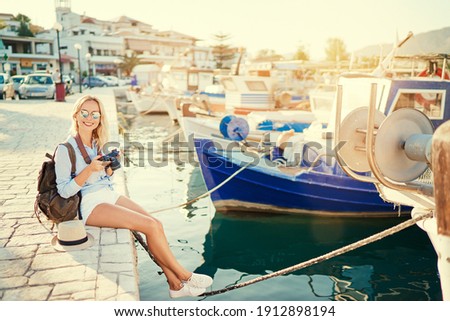 Photography and travel. Young woman with rucksack holding camera sitting on wharf with beautiful sea view with fishing boats.