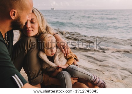 Mother, dad and baby daughter next to the sea smiling. Baby girl with blonde hair, in a warm fur vest. Spring time, sun is shining. Travel abroad. Happy young family. 