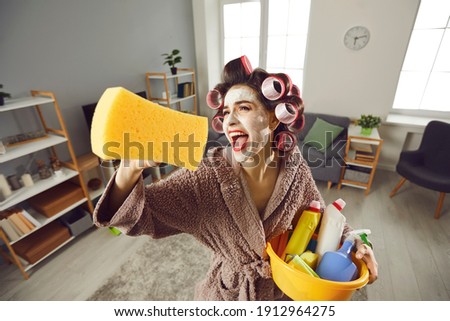 Tired desperate housewife holding yellow sponge and screaming like crazy while cleaning house. Funny young woman in hair rollers and beauty skin care face mask tidying up home, singing and having fun Royalty-Free Stock Photo #1912964275