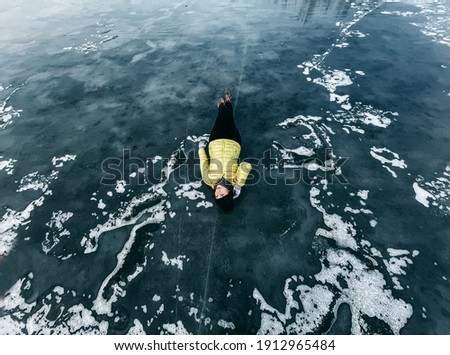 Portrait of a beautiful young European woman in a yellow jacket lies on a blue ice-frozen lake with snow and cracks in winter. Looks like a planet, clouds, water, abstraction. Nature, cold, travel