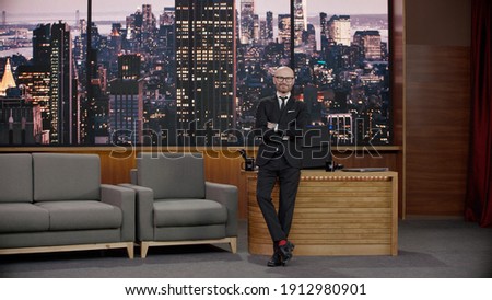 Late-night talk show host is leaning against his table in studio, looking into camera. TV broadcast style show Royalty-Free Stock Photo #1912980901
