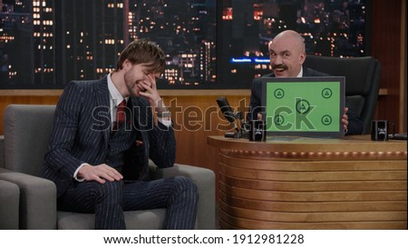 Late-night talk show host showing a green board with tracking points to celebrity guest in a studio. TV broadcast style show