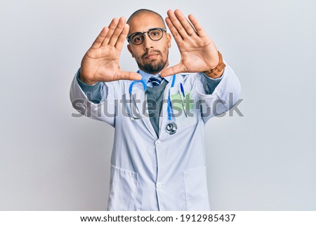 Hispanic adult man wearing doctor uniform and stethoscope doing frame using hands palms and fingers, camera perspective 