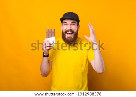 Portrait of cheerful young bearded hipster man holding bar of chocolate over yellow background.