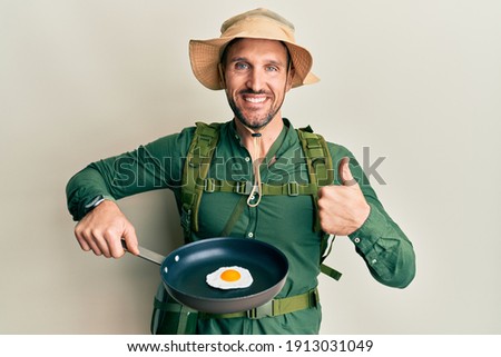 Handsome man with beard wearing explorer hat cooking fried egg smiling happy and positive, thumb up doing excellent and approval sign 