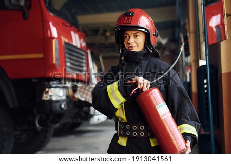 Holds extinguisher in hands. Female firefighter in protective uniform standing near truck.