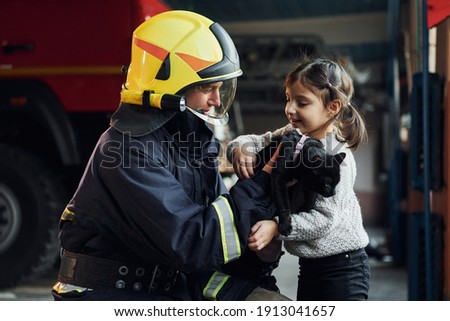Cute black cat. Happy little girl is with male firefighter in protective uniform.