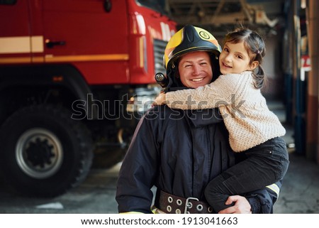 Happy little girl is with male firefighter in protective uniform.
