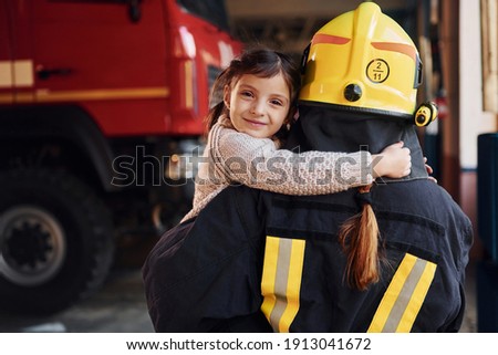 Happy little girl is with female firefighter in protective uniform.