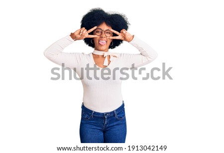 Young african american girl wearing casual clothes and glasses doing peace symbol with fingers over face, smiling cheerful showing victory 