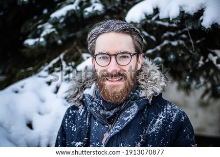 Young bearded man in winter clothes sprinkled in snow, looking at the camera and smiling while walking in snow on a winter sunny day. Winter fun, throwing snowballs, outdoor recreation.