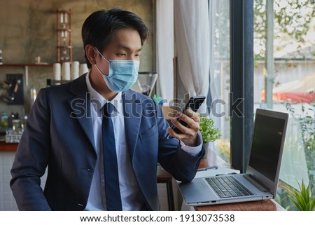 Front Right Asian Businessman in Suit Wear Face Mask Using Smartphone and Laptop in Coffee Shop Scene. Asian Businessman Work from Anywhere with Technology in Covid 19 Situation