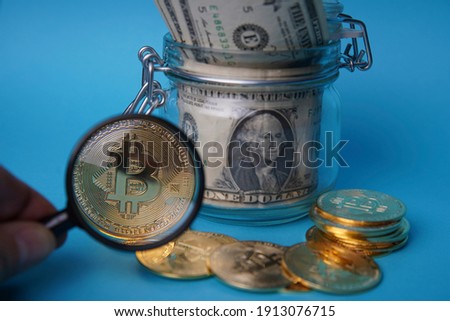 A glass jar full of dollars viewed through a magnifying glass. An easy way to save real money. Next to it are several gold bitcoin digital cryptocurrency coins. Blue background. Bank image and photo.