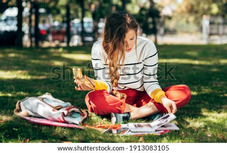 Horizontal image of a student female sitting on the green grass at the college campus on a sunny day, have lunch and studying. The hungry young woman takes a rest learning and eating in the park. Royalty-Free Stock Photo #1913083105