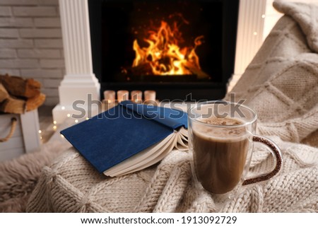 Cup of coffee and book on plaid near fireplace indoors. Cozy atmosphere
