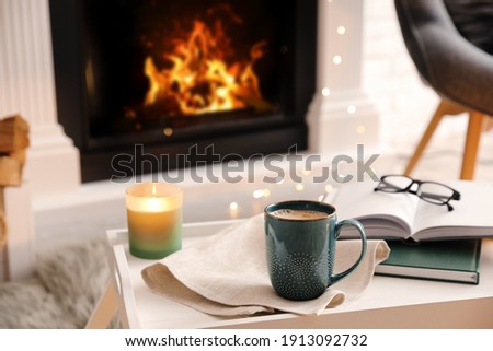 Cup of coffee, burning candle and books on tray near fireplace indoors. Cozy atmosphere Royalty-Free Stock Photo #1913092732