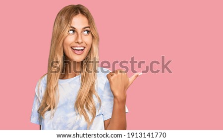 Beautiful blonde young woman wearing casual tie dye shirt smiling with happy face looking and pointing to the side with thumb up.  Royalty-Free Stock Photo #1913141770