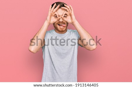 Young redhead man wearing casual grey t shirt doing ok gesture like binoculars sticking tongue out, eyes looking through fingers. crazy expression. 