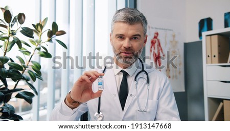 Close up portrait of happy handsome Caucasian middle-aged experienced male doctor immunologist looking at camera and speaking on video call with patient in cabinet in hospital, showing covid vaccines Royalty-Free Stock Photo #1913147668