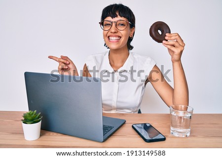 Beautiful brunettte woman working at the office eating chocolate doughnut smiling happy pointing with hand and finger to the side 