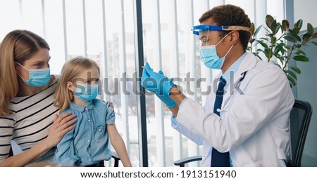 Portrait of African American young handsome happy man doctor specialist immunologist in mask injecting cute Caucasian small kid girl sitting in clinic with mom, disease prevention, vaccination concept Royalty-Free Stock Photo #1913151940