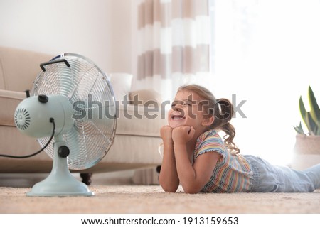 Little girl enjoying air flow from fan on floor in living room. Summer heat Royalty-Free Stock Photo #1913159653