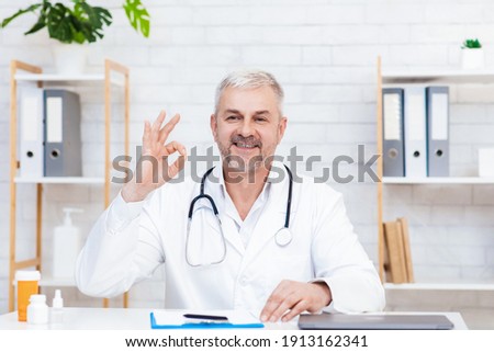 Doctor working exam, health care and medical and test results. Smiling senior man in white coat gesturing, shows sign ok with hand and advices to patient at video call, sits at desk with medicines