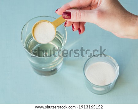 Glass with collagen dissolved in water and collagen protein powder on white wooden table. Female hand holds a measure scoop. Healthy lifestyle concept.