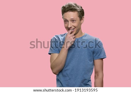 Smiling european boy is touching his lips with his finger. Schoolboy in t-shirt isolated on pink background.