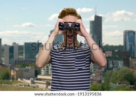 Shocked young teen boy looks through binoculars. Urban cityscape on the background.