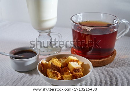 Square toasted pieces of homemade delicious rusk, hardtack, Dryasdust, zwieback, Liquid honey in a saucer, glass of milk and black tee in a cap on a white tablecloth