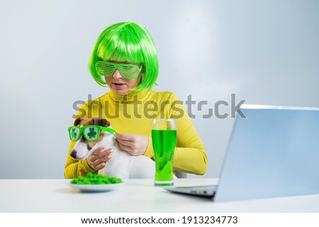 A young woman in a green wig and cheerful glasses drinks beer and bites glazed nuts. A girl sits with a dog at a table and celebrates st patrick's day online chatting with friends on a laptop.